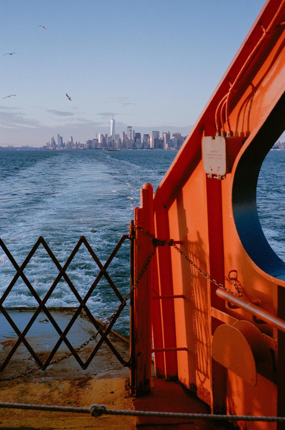 A ferry departs from New York City, leaving a wake in the water as seagulls fly overhead.