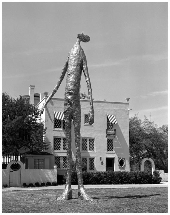 A tall, thin, metallic sculpture of a figure stands in a grassy area in front of a building with striped awnings.