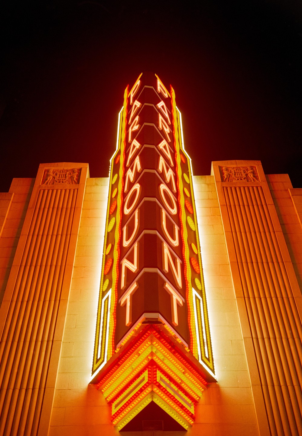 A tall, illuminated sign for "Paramount" stands against a dark night sky, with architectural details of the building visible on either side.