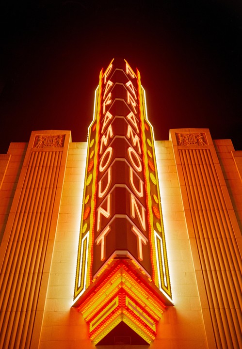 A tall, illuminated sign for "Paramount" stands against a dark night sky, with architectural details of the building visible on either side.