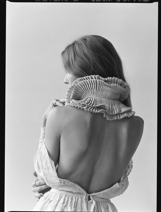 A woman with long hair and a ruffled collar is seen from behind, with her back exposed.