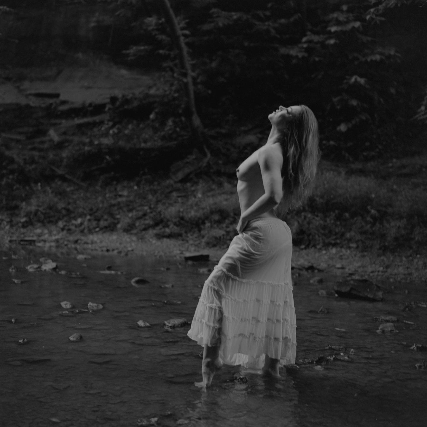 A topless woman in a long skirt stands in a shallow stream, looking up with her hair flowing back.