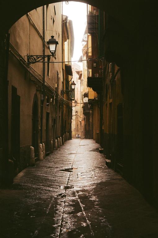 A narrow, wet alleyway in Florence is illuminated by light at the far end and from a streetlamp on the left.