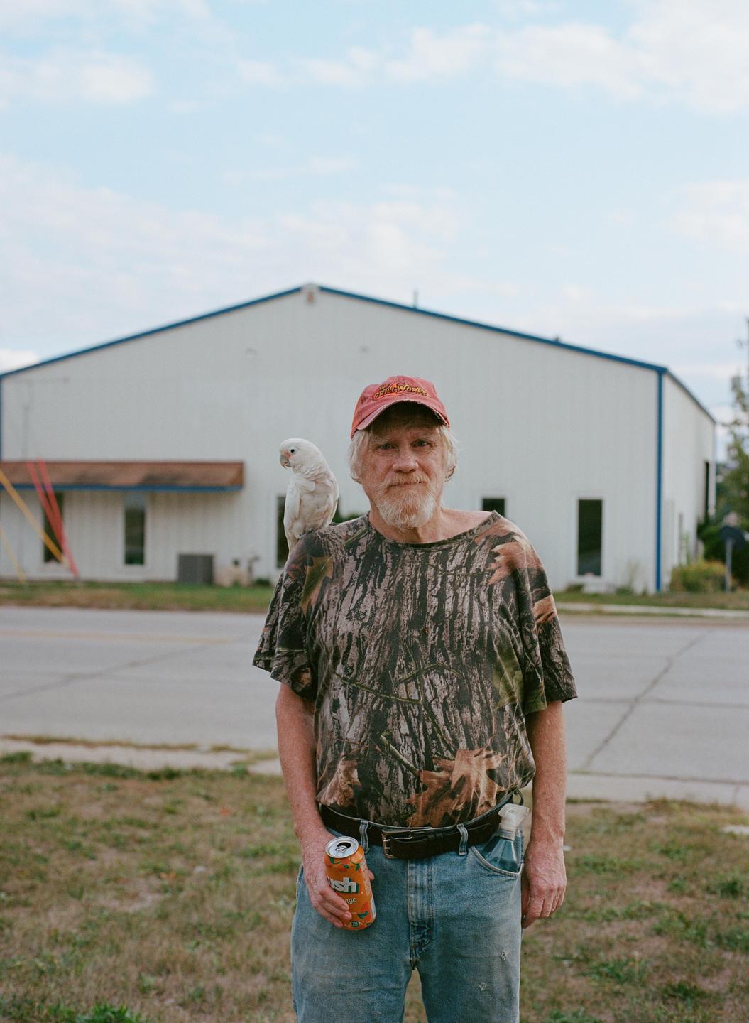 A man with a beard and a parrot on his shoulder stands outdoors holding a soda can.
