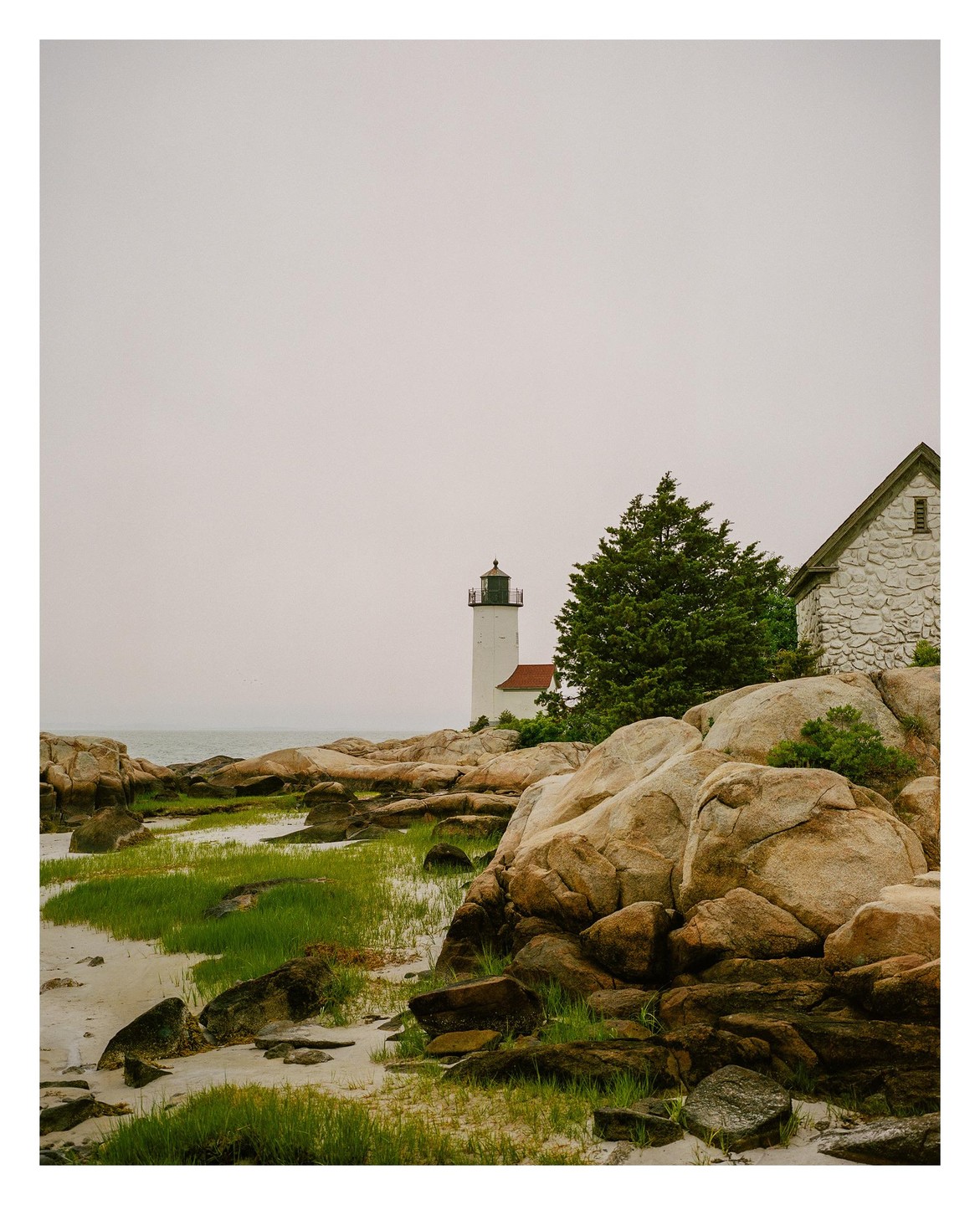 A lighthouse stands on a rocky shore with green grass and a stone building under a hazy sky.