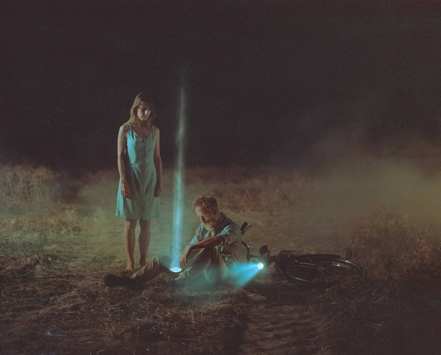 A woman stands barefoot next to a man sitting on the ground with a bicycle and a flashlight, all illuminated by a mysterious light beam in a dark, hazy field.