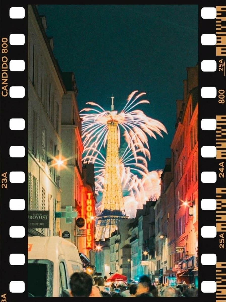 Fireworks explode over the Eiffel Tower, viewed from a crowded street lined with buildings at night.