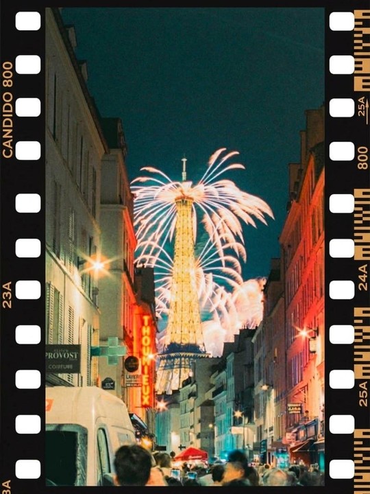 Fireworks explode over the Eiffel Tower, viewed from a crowded street lined with buildings at night.