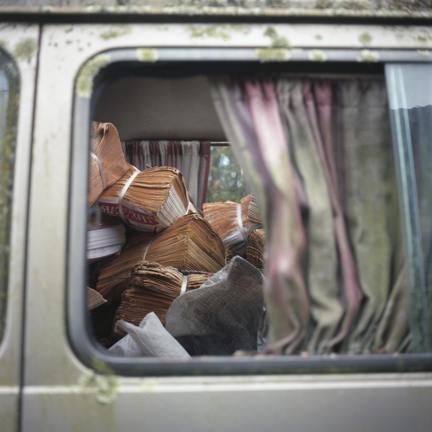 Bundles of paper are stacked inside a vehicle with curtains on the windows.
