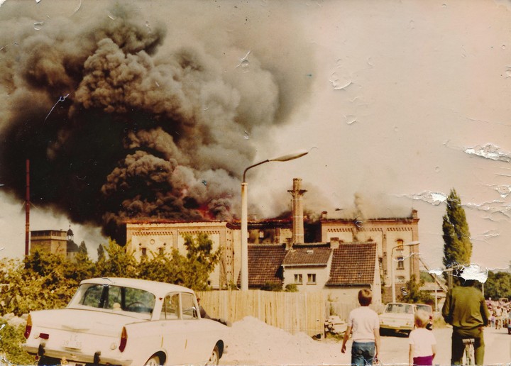 A large building is on fire, emitting thick black smoke into the sky, as people watch from the street.
