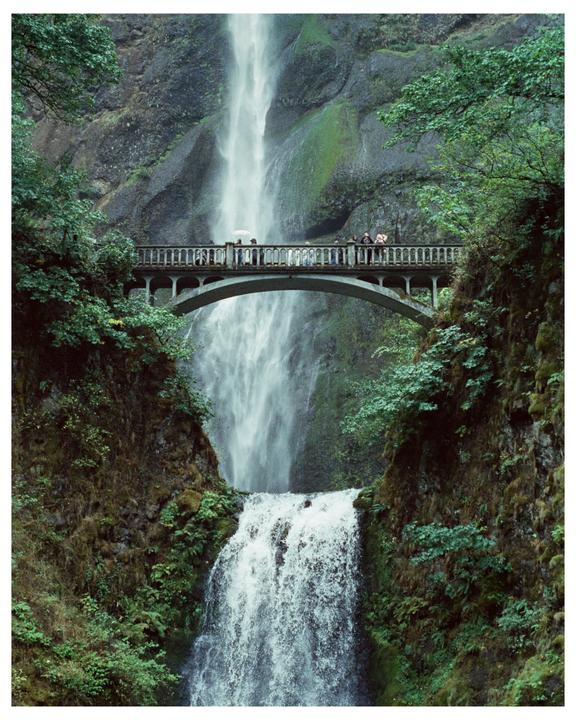 A tall waterfall cascades down a rocky cliff, with a stone bridge spanning the gorge in front of it, where several people are standing.