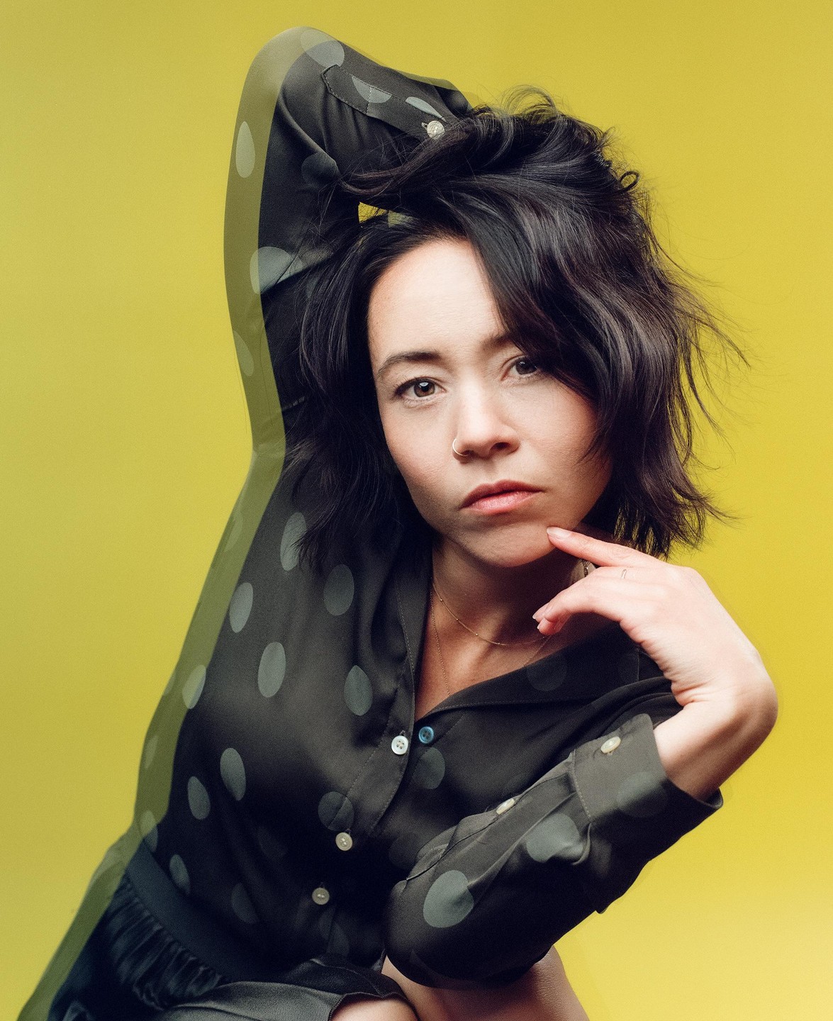 A woman with dark hair and a polka dot shirt poses against a yellow background.