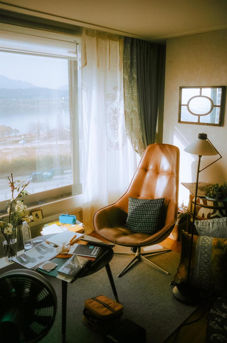 Sunlight streams into a cozy reading nook with a brown leather chair, a small table with books, and a window overlooking a body of water and mountains.