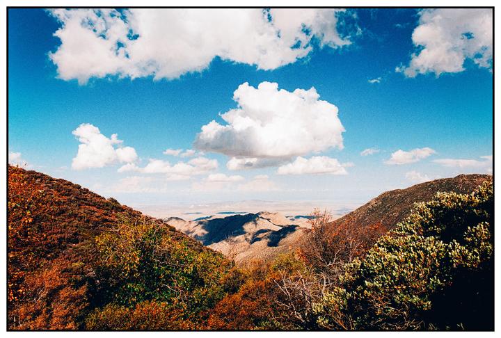 A wide landscape shows a valley with mountains and hills under a blue sky with white clouds.