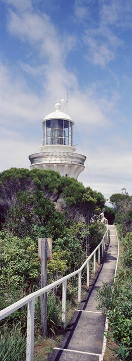 A white lighthouse with a glass top stands above green bushes, with a paved path and white railing leading towards it under a blue sky.