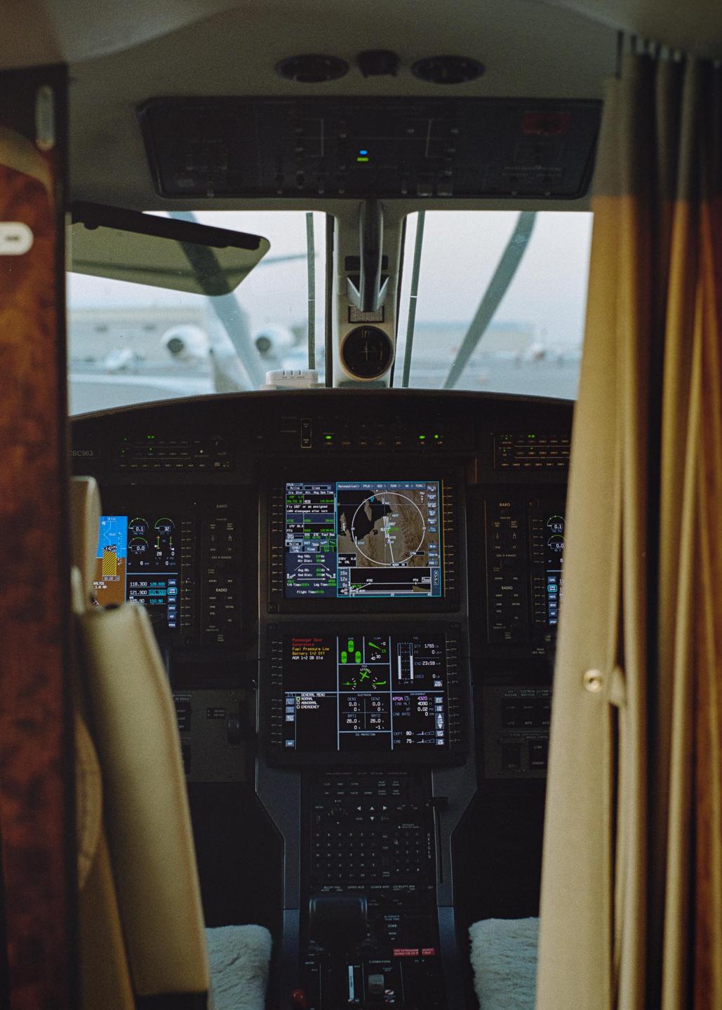 The cockpit of an airplane is visible with multiple screens displaying flight information and a view of other aircraft on the tarmac through the windshield.