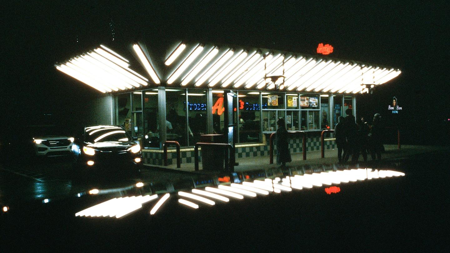 A brightly lit fast food restaurant with several people standing outside is visible at night.