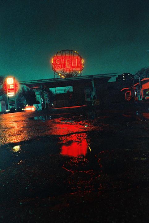 A Shell gas station with a prominent neon sign is illuminated at night, reflecting in puddles on the wet ground.