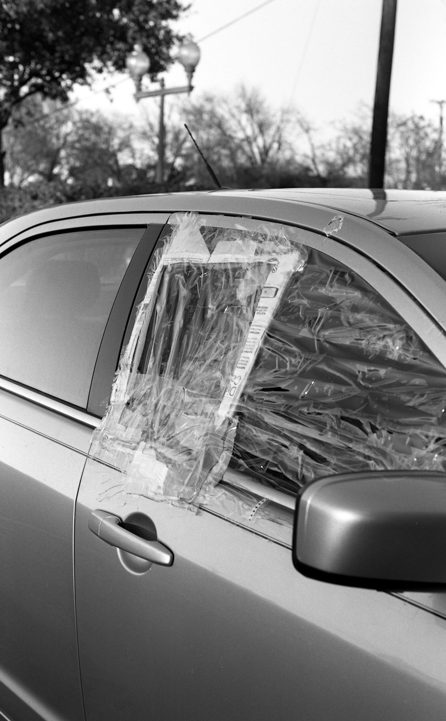 A car with a broken window covered in plastic and tape is parked on a street.