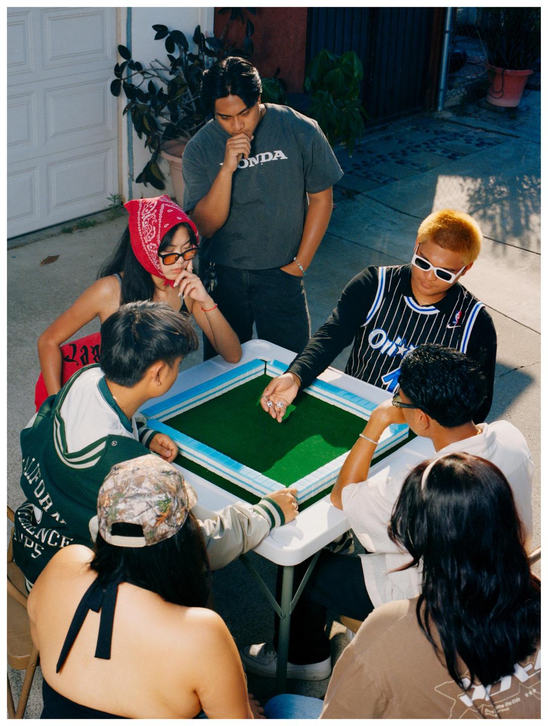 A group of young adults are gathered around a table playing mahjong outdoors.