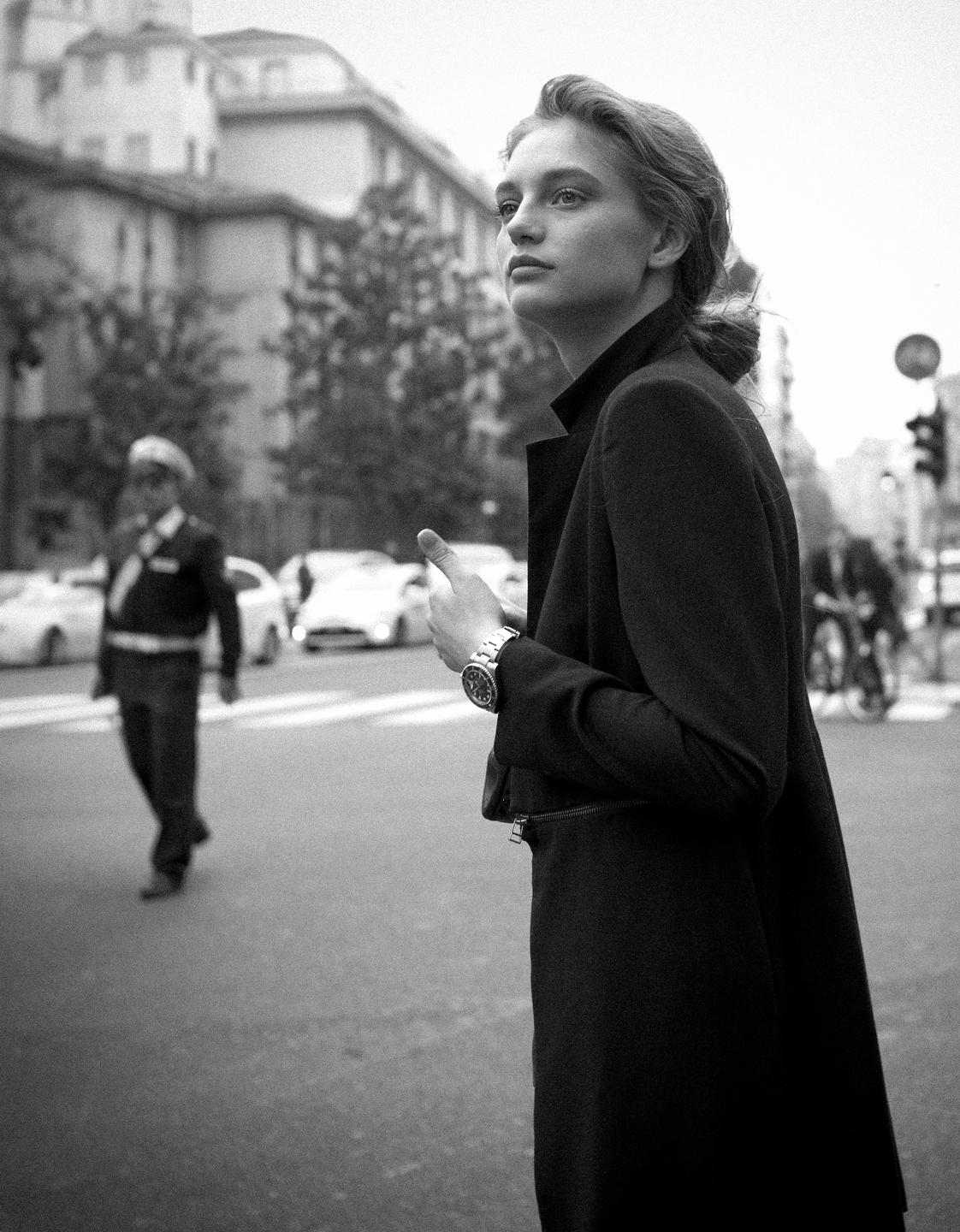 A woman in a dark coat stands on a city street with a man in uniform in the background.