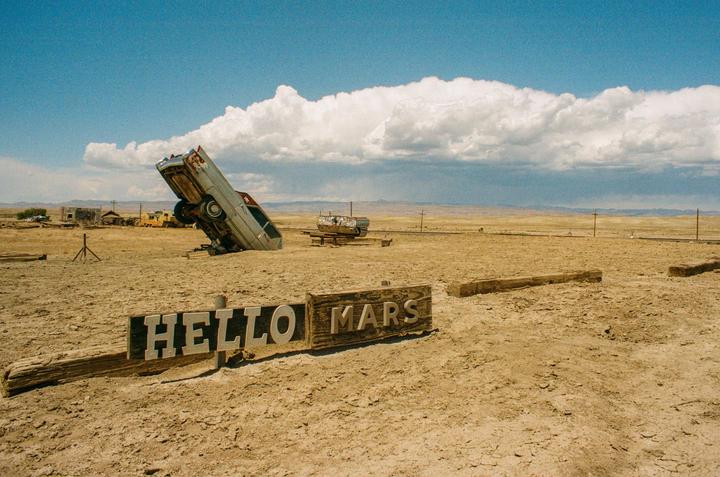 A sign reading "HELLO MARS" is in the foreground of a desert landscape with an old car buried nose-down in the sand.