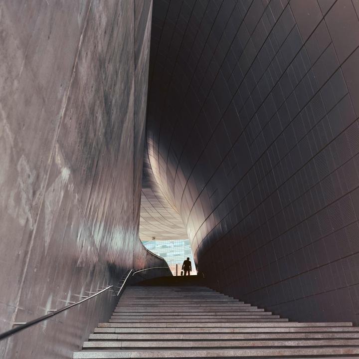 A man stands at the top of a long staircase between two curving modern walls.