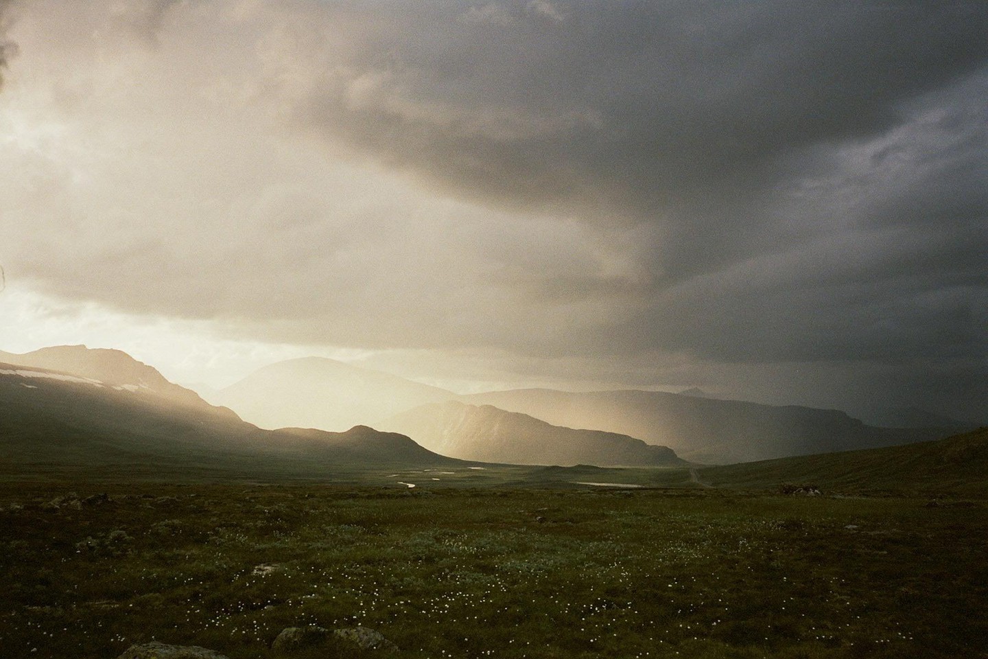 Sunlight breaks through dark clouds, illuminating a vast green valley with distant mountains and a winding river.