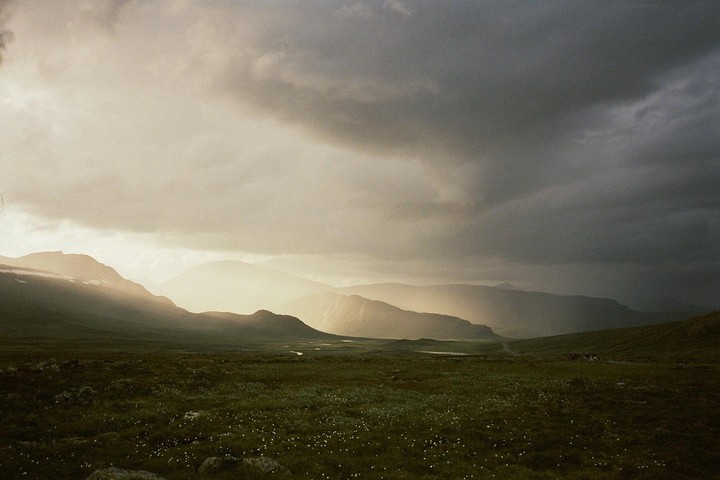 Sunlight breaks through dark clouds, illuminating a vast green valley with distant mountains and a winding river.