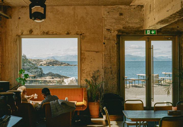 A couple sits at a table inside a cafe with a view of the ocean and rocky coastline through a large window and glass door.