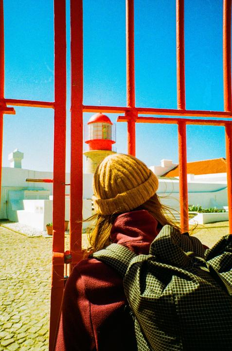 A person wearing a beanie and backpack looks through red bars at a lighthouse under a clear blue sky.