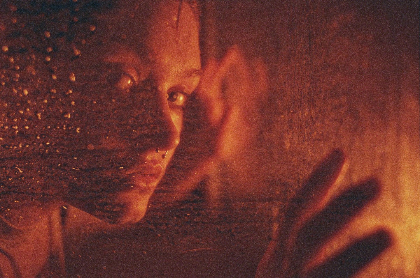 A woman's face and hand are visible through a wet surface, with warm light illuminating the scene.