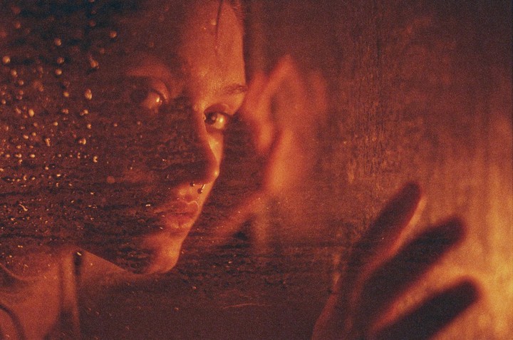 A woman's face and hand are visible through a wet surface, with warm light illuminating the scene.