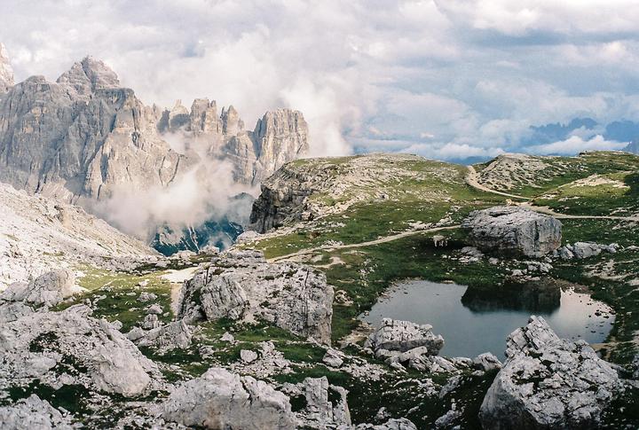 A mountain landscape shows a small lake surrounded by rocks and green grass, with a path leading up to a mountain range partially obscured by clouds.