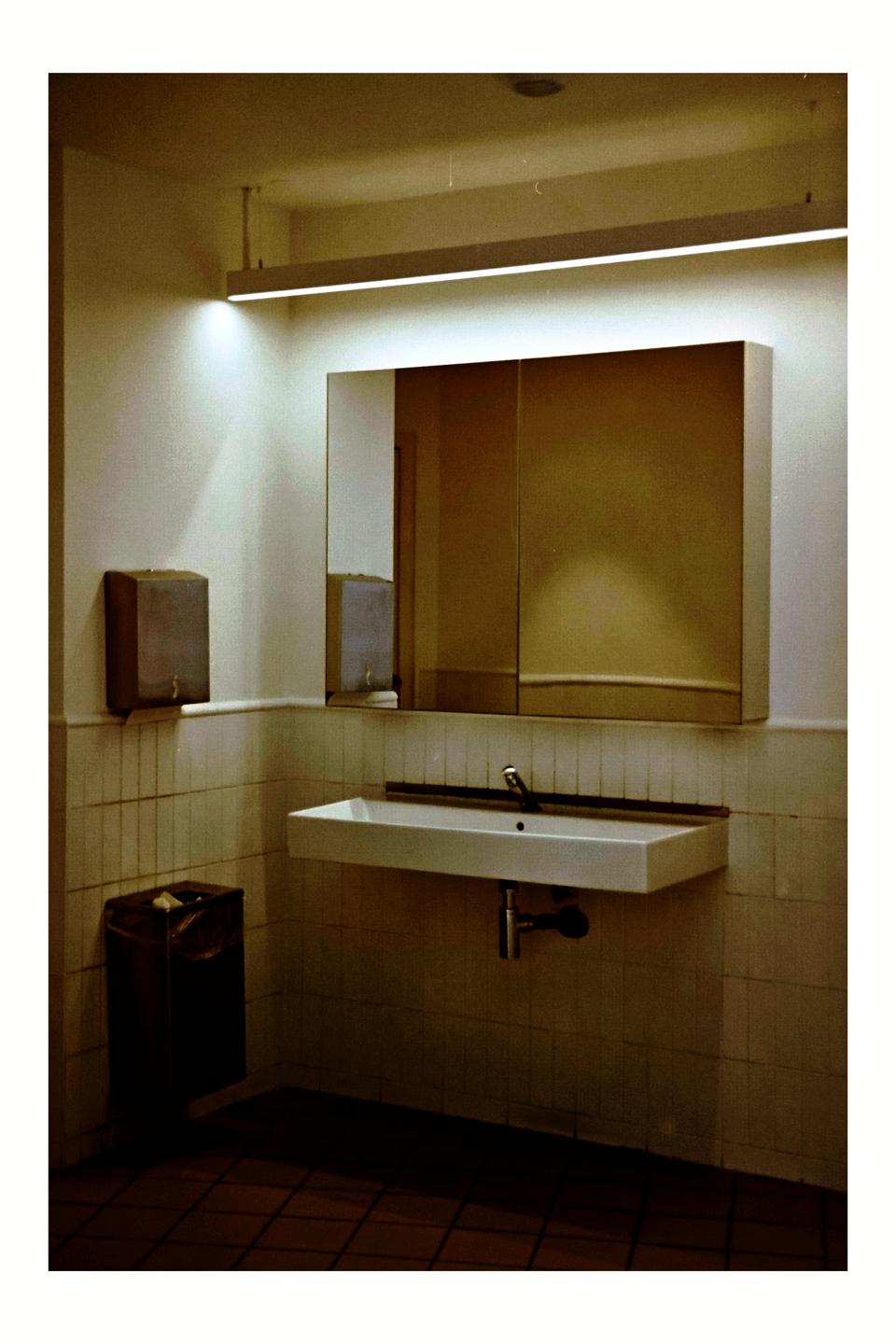A sink, mirror, and trash can are visible in a public restroom.