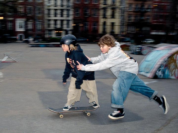 A boy in a helmet is on a skateboard while another boy pushes him from behind in a skatepark.