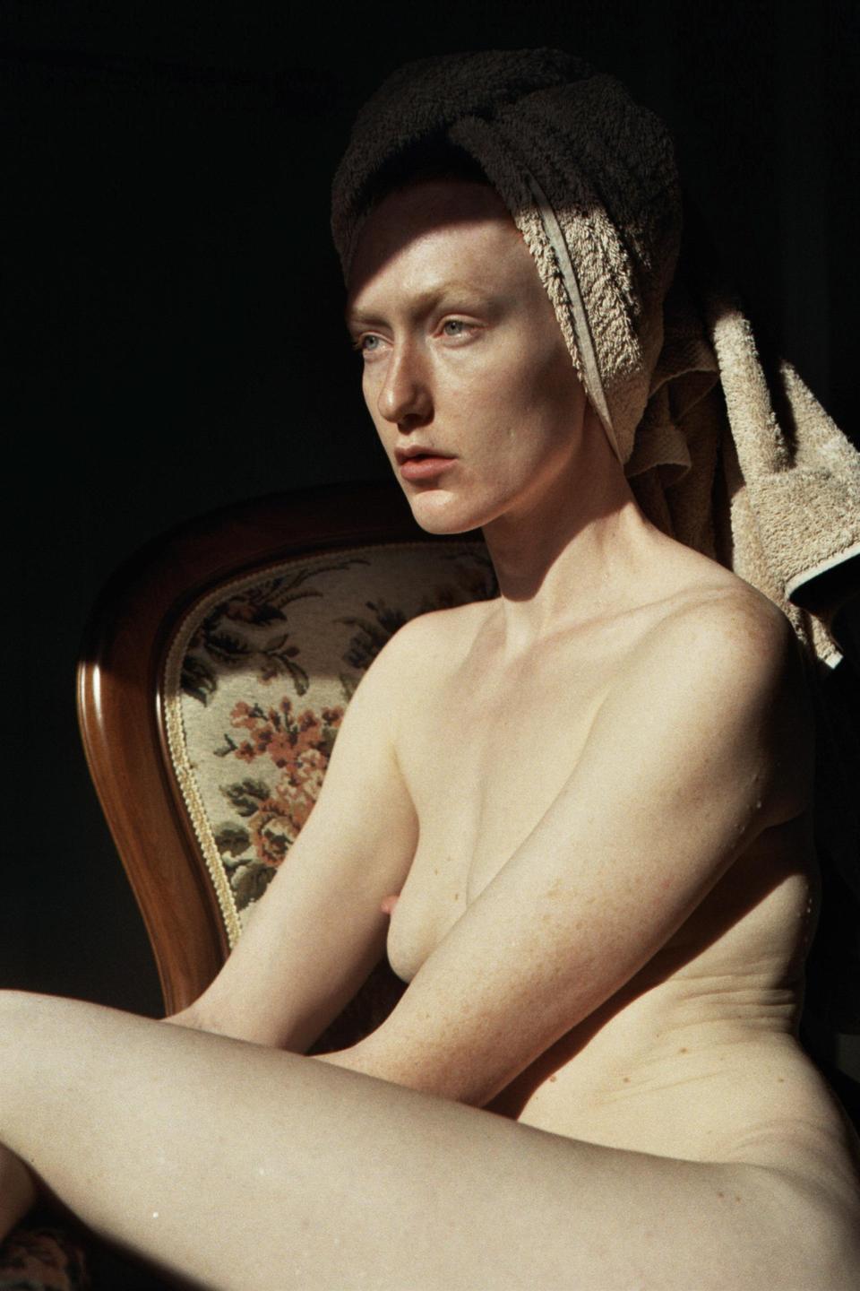 A woman with a towel wrapped around her head sits in a chair, illuminated by a shaft of light.