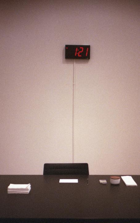 A digital clock displaying "1:21" hangs above a dark table with a chair and some items.