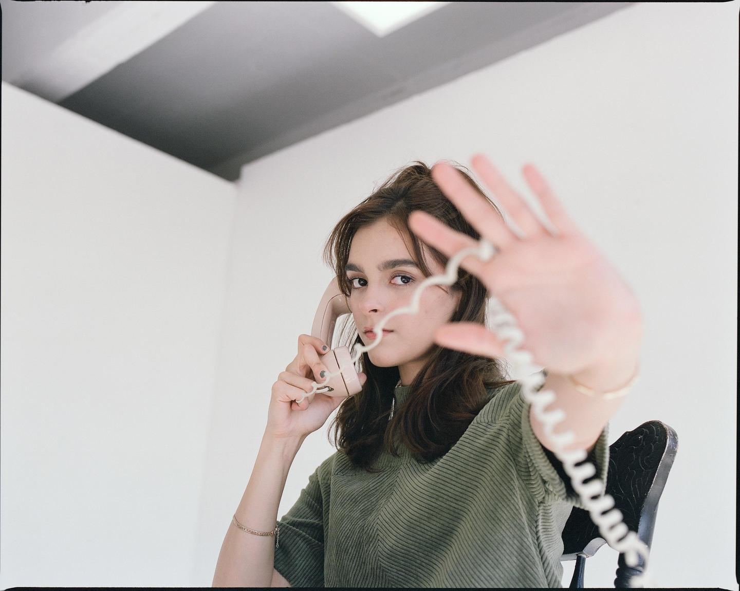 A woman holds a telephone receiver to her ear while extending her other hand toward the viewer, with the telephone cord in the foreground.