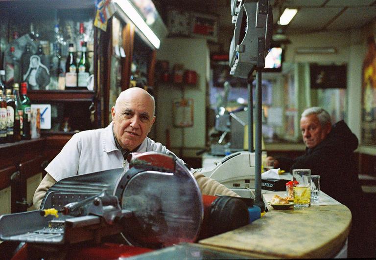A man in a white shirt stands behind a deli counter with a meat slicer, looking at the viewer, while another man sits at the counter in the background.