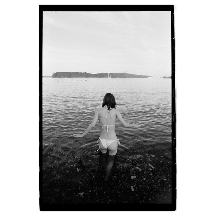 A woman in a bikini stands in shallow water looking out at a body of water with an island in the distance.
