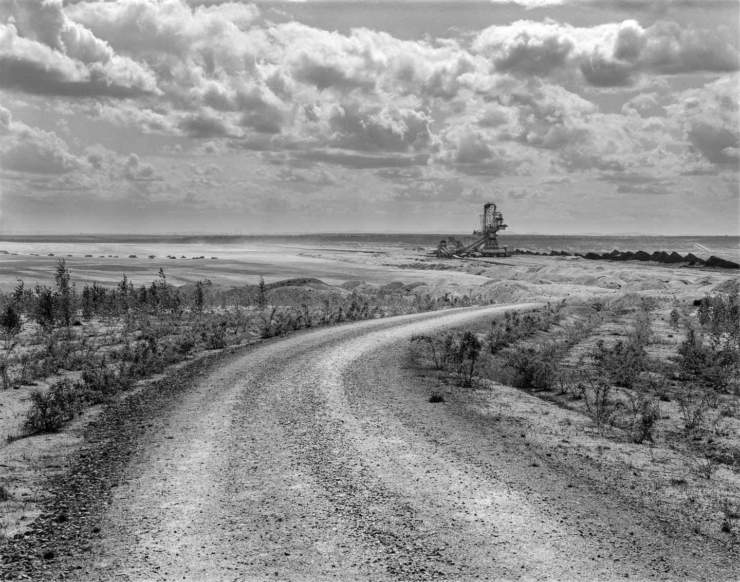 A dirt road curves through a barren landscape with a large mining machine in the distance under a cloudy sky.