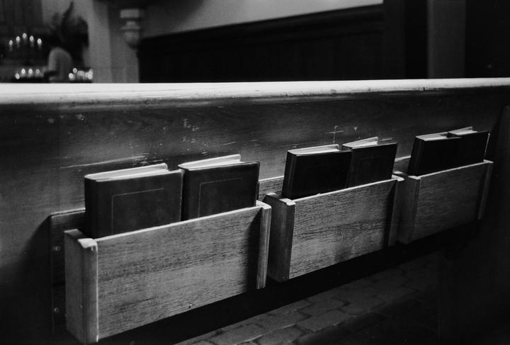 Several books are stored in wooden holders on the back of a church pew.