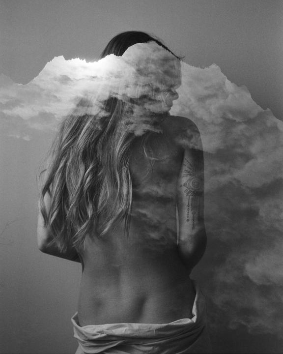 A woman with long hair is seen from behind in a double exposure with clouds.