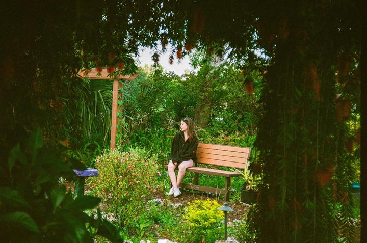 A woman sits on a wooden bench in a lush garden, framed by dark foliage.