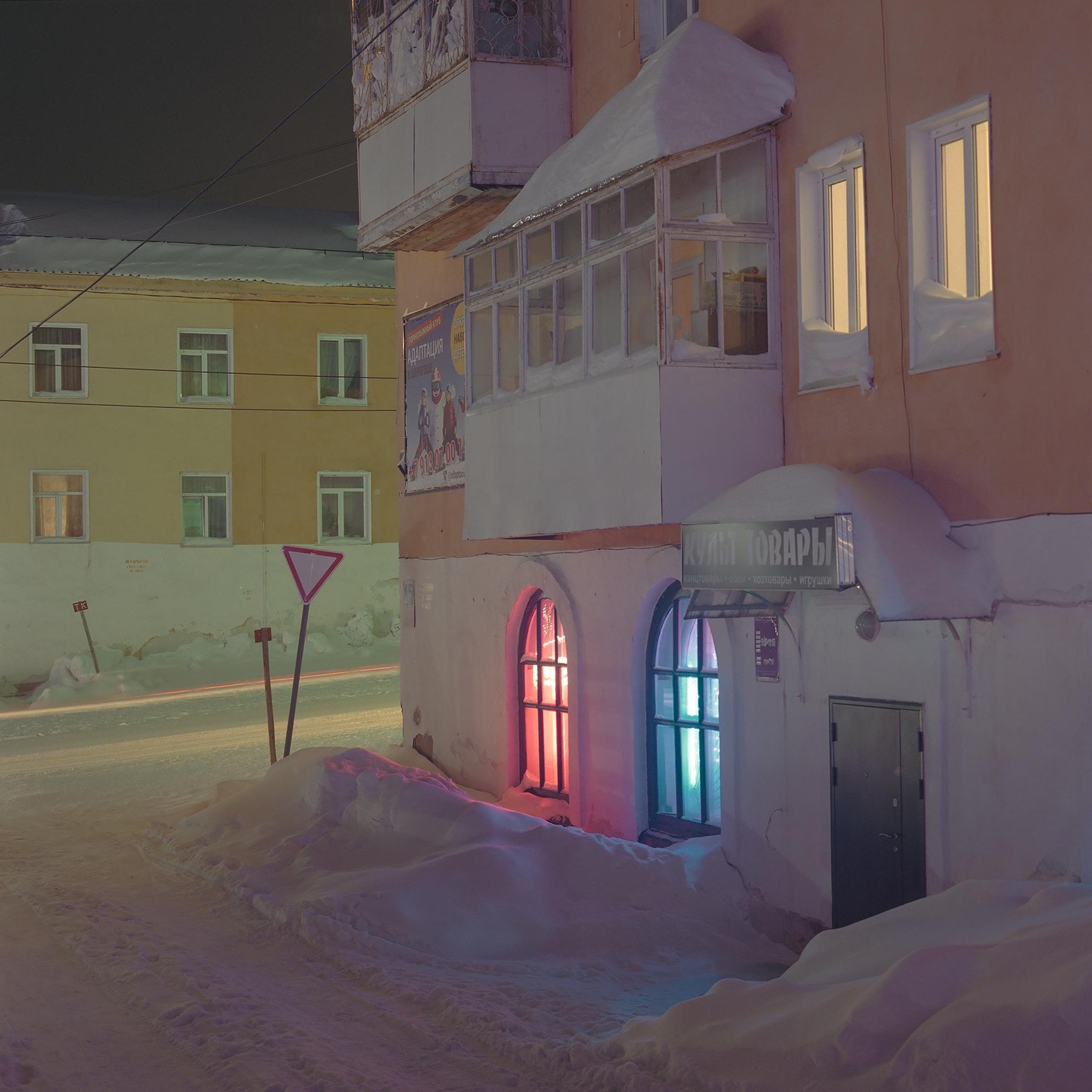 A snow-covered street at night is lined with buildings, one of which has windows glowing red and blue.