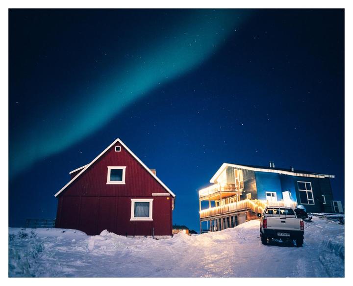 A green aurora borealis streaks across a dark night sky above two houses and a pickup truck in a snowy landscape.
