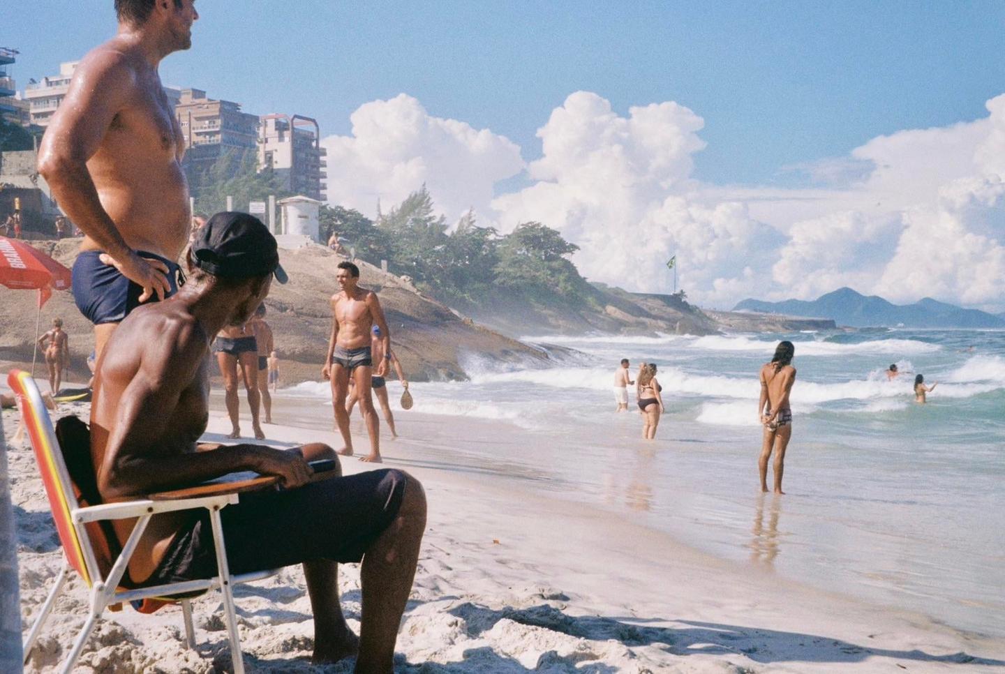 A group of people are enjoying a sunny day at a beach with waves crashing on the shore and buildings on a hill in the background.