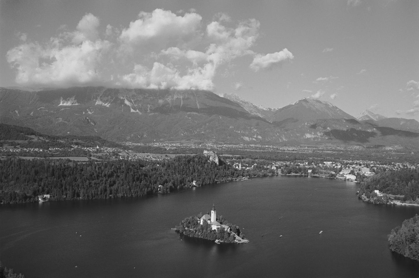 A small island with a church and bell tower sits in a lake surrounded by a town, forest, and mountains under a cloudy sky.