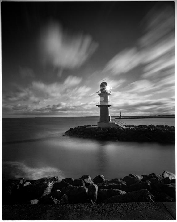 A lighthouse stands on a rocky jetty with blurred clouds in the sky and smooth water below.
